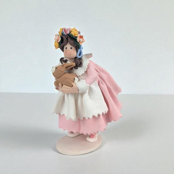 Handmade polymer clay figurine of a girl with a floral crown, wearing a pink dress and white scalloped apron while holding a brown bunny. The texture is matte and dough-like with soft pastel tones and a folk-art silhouette.