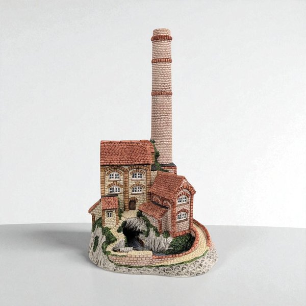 A detailed architectural miniature of a multi-story stone mill featuring a towering, slender brick chimney. The piece displays a textured cream and tan facade, terracotta-colored gabled roofs, and intricate arched windows set against a rocky, moss-covered base.