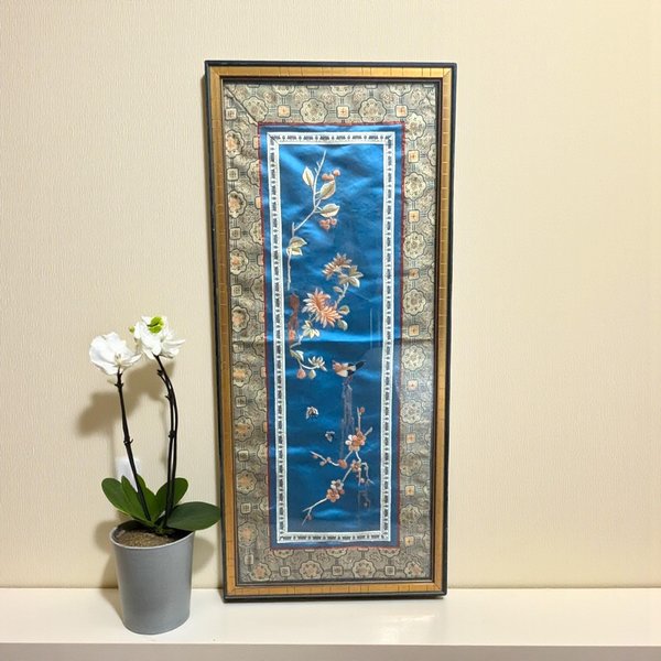 Vertical rectangular framed textile. A central blue silk panel features embroidered flora, berries, and a small bird. The panel is bordered by a wide cream and gold jacquard fabric with floral medallions, set within a slim dark frame with a gold inner lip.