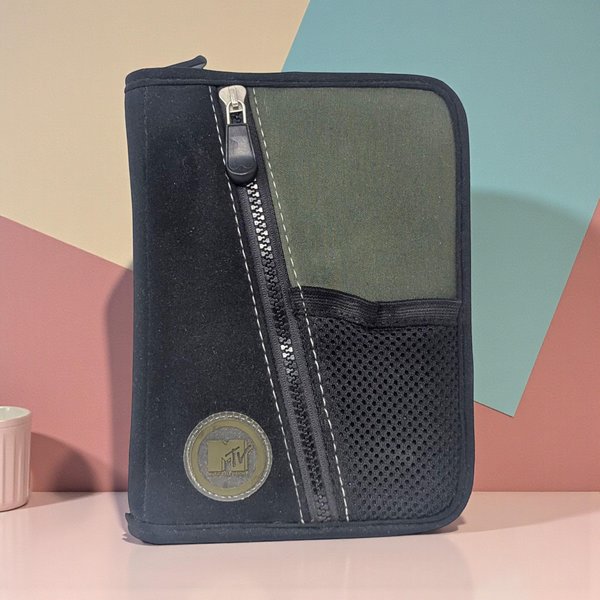 A black and olive green zippered binder featuring a circular MTV logo. The exterior has a rugged canvas texture with a decorative diagonal zipper, white contrast stitching, and a small black mesh pocket. The silhouette is rectangular with rounded corners.