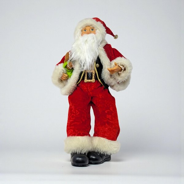 A seated Santa figure in an elf-style pose, wearing a red velvet suit with green trim and a tall pointed hat. He holds a stuffed gingerbread man and features long, dangling legs with pointed black boots.