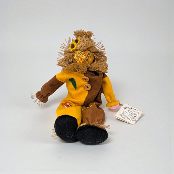A seated scarecrow figure wearing a denim shirt, plaid vest, and a tall burlap hat with a sunflower. The figure has straw-like hair and hands, with long, limp legs designed to hang over an edge.