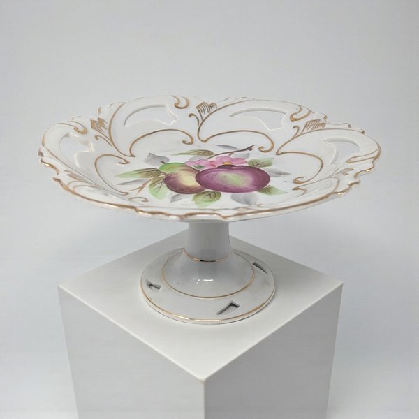 A white porcelain pedestal dish featuring a hand-painted plum and pear motif at the center. The shallow bowl has a decorative pierced or reticulated rim accented with gold filigree and a matching footed base.