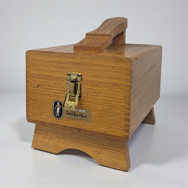 A light-toned oak shoe shine box featuring a built-in footrest on top. The chest has finger-jointed corners, a brass-tone metal latch, and an original Esquire Shoe Care Chest label on the front.