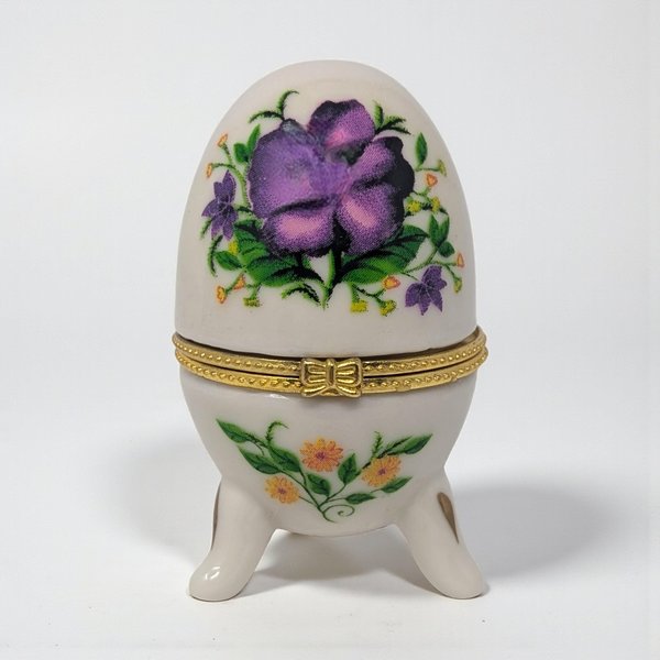 A small, egg-shaped white ceramic box with a hinged gold metal closure. The exterior is decorated with delicate purple violet sprays and green leaves on both the lid and base.