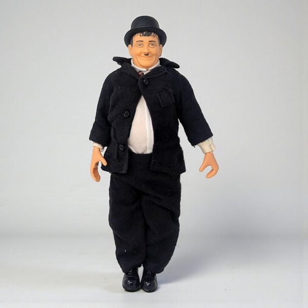 A vinyl character doll of comedian Oliver Hardy featuring his iconic bowler hat, black suit, and mustache. The figure has a rounded physique and visible joints at the elbows and knees.