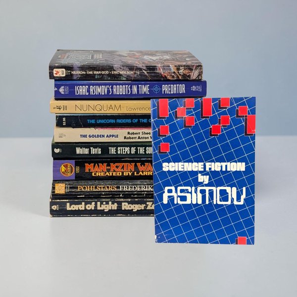 A stack of nine vintage mass-market paperbacks sits vertically against a neutral background, with a tenth book, Science Fiction by Asimov, leaning against the front. The spines show titles from authors like Roger Zelazny and Frederik Pohl. The front book features a vibrant blue grid pattern with red geometric squares.