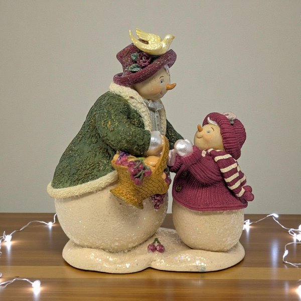 Matte resin figurine of two women in winter coats and hats walking side-by-side. One wears a green coat with a red scarf, the other a red coat with a red scarf.