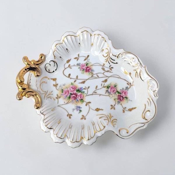 A low, oval porcelain bowl with a multi-colored floral bouquet in the center. The texture is a smooth, high-gloss white ceramic with a scalloped edge and a metallic gold trim.