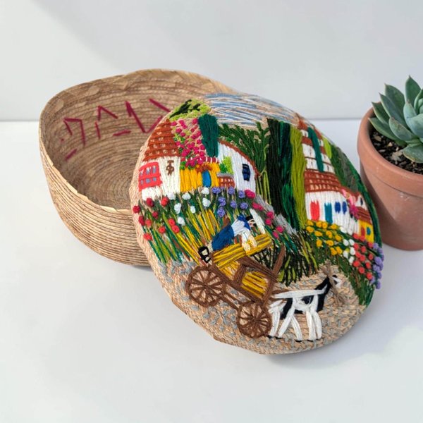 A round woven wicker bowl with a matching lid featuring a colorful embroidered floral design. The texture is a mix of coarse natural fibers and smooth, raised threadwork in a squat, cylindrical silhouette.