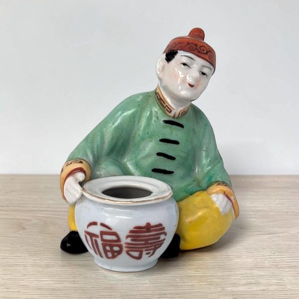 A porcelain figurine of a seated person in a green robe and red cap leaning over a small white pot. The pot features red calligraphic characters, and the figure is set against a plain background on a light wood surface.