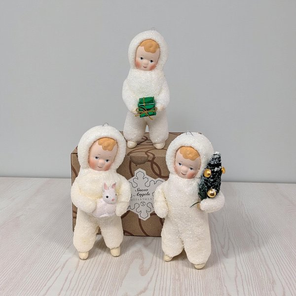 A set of three ceramic figurines depicting children in white winter coats and hats, posed as if making snow angels. They feature a soft matte finish with iridescent glitter accents, and are shown alongside their original Department 56 boxes.