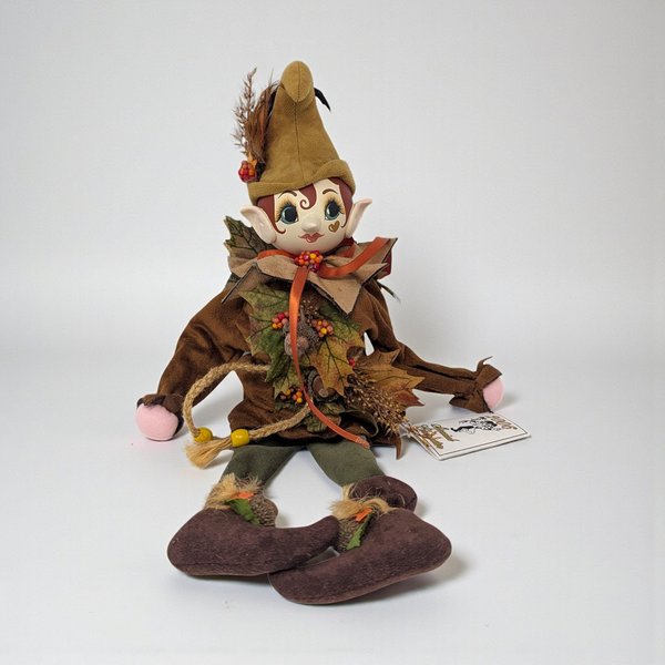 A seated woodland nymph doll with a hand-carved wooden face and soft, fabric hands. She wears a rustic forest outfit in mossy green and brown tones, featuring long, unfilled legs designed to drape over a shelf edge.