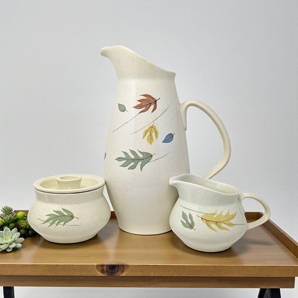 A three-piece ceramic service set featuring a tall, elegant pitcher and matching cream and sugar vessels. The set is finished in a warm tan speckled glaze and decorated with vibrant autumnal leaf motifs in shades of orange, yellow, and brown.