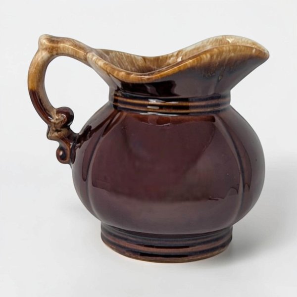 A squat, round ceramic pitcher with a wide pouring spout and a large integrated handle. It features a glossy dark brown glaze with a thick, frothy cream-colored drip effect around the top rim.