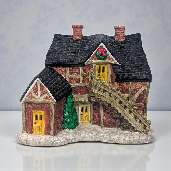 A snow-dusted ceramic village house featuring a steeply pitched roof, handpainted green shutters, and a bright red front door. Small cut-out windows allow light to glow from within the cream-colored structure.