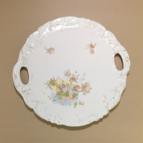 A round white ceramic serving plate with two integrated tab handles. The center features a large multi-colored floral spray, and the scalloped rim shows aged gold gilt accents with visible wear.