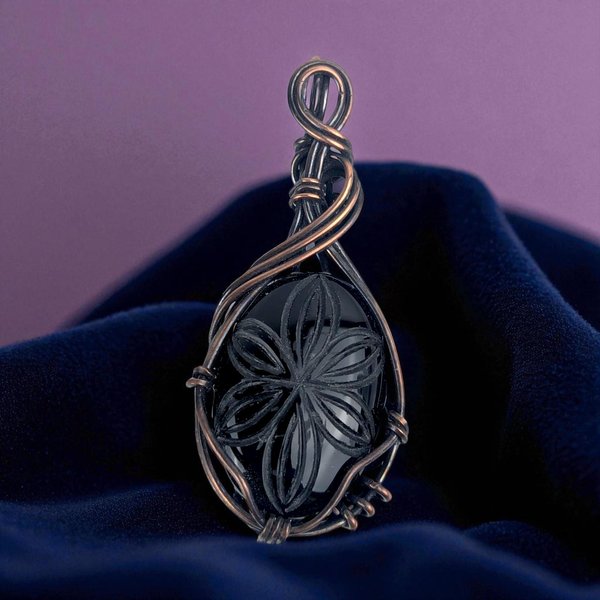 A teardrop-shaped black onyx stone with a deeply carved floral pattern, encased in dense oxidized copper wire weaving. The metal has a dark, antiqued patina with intricate coiled and layered textures.
