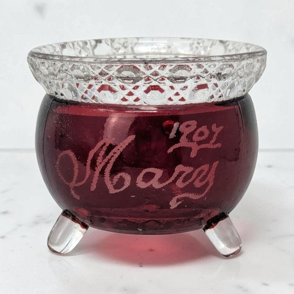 A small, three-footed glass kettle with a deep ruby red body and clear, diamond-cut rim. The red surface features an etched cursive name, Mary, and the year 1907. The texture is smooth glass contrasted by the textured, geometric collar.