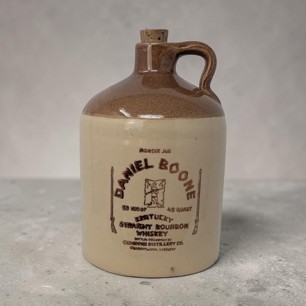 A 7-inch 1974 Daniel Boone bourbon stoneware jug. Features a two-tone cream base and tobacco-brown glazed shoulder with a black graphic of Daniel Boone in a coonskin cap. The cylindrical silhouette includes a finger loop handle and an original cork.