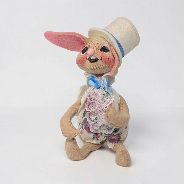 Tan felt bunny wearing a white top hat with a blue ribbon and a white floral vest. He features a lace ruffle at his neck and a tiny blue bow. One long ear is lined in pink felt and he has a toothy hand-painted grin.
